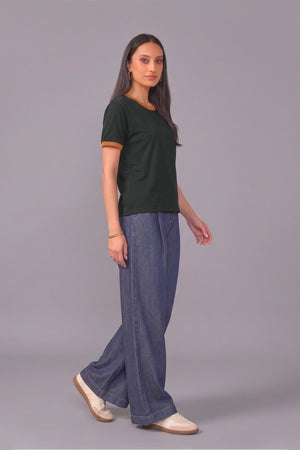 A woman modeling a dark green ringer t-shirt with tan trim and wide-leg blue jeans, standing against a solid gray studio background.