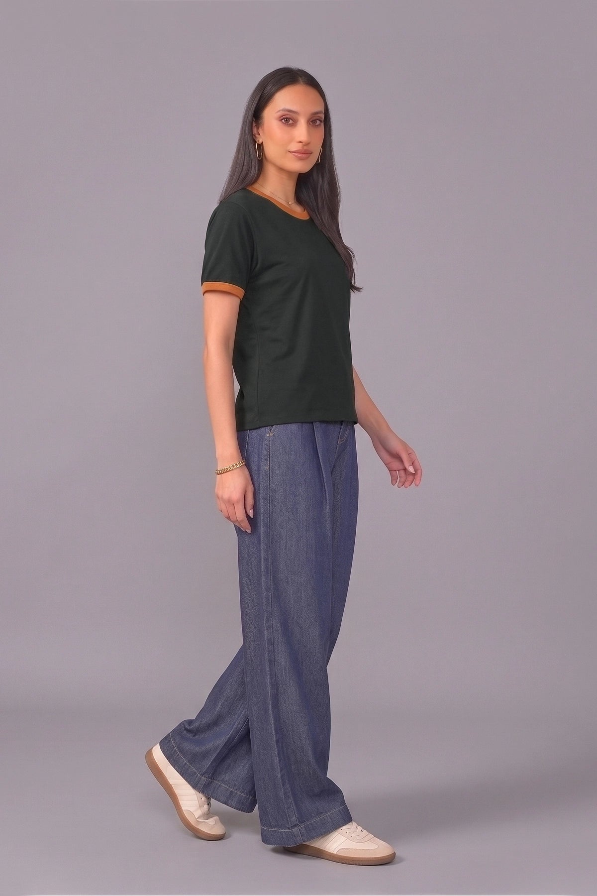 A woman modeling a dark green ringer t-shirt with tan trim and wide-leg blue jeans, standing against a solid gray studio background.